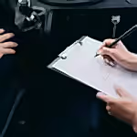 Close-up of a person writing on a clipboard inside a car, showing hands and a gear shift.